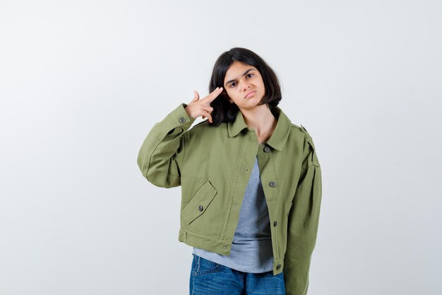 Little girl shouting herself with hand gun in coat, t-shirt, jeans and looking sorrowful. front view.