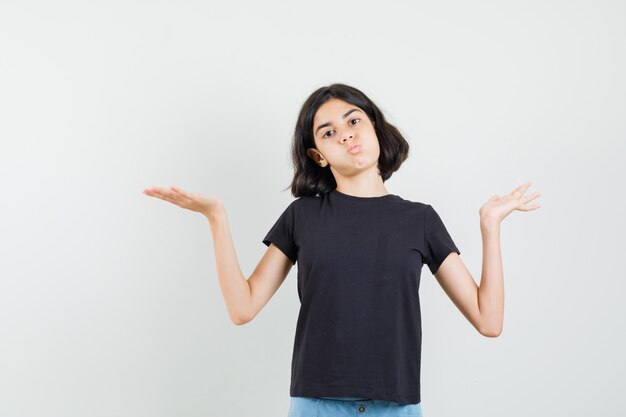 Little girl showing helpless gesture by shrugging in black t-shirt, shorts and looking confused , front view.