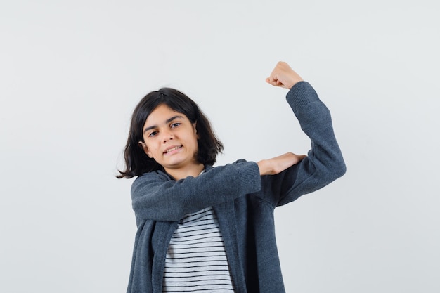 Маленькая девочка показывает мышцы руки в футболке, куртке и выглядит сильной