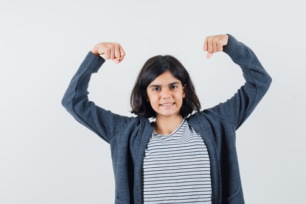 Little girl showing muscles of arms in t-shirt, jacket and looking powerful.