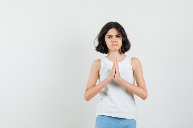 Little girl showing namaste gesture in white blouse, shorts and looking gloomy , front view.