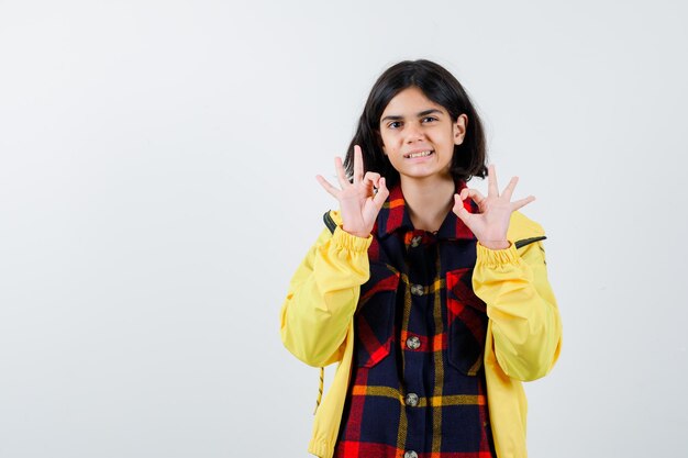 Little girl showing ok gesture in checked shirt, jacket and looking happy. front view.