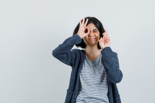 Little girl showing ok gesture, keeping fingers crossed in t-shirt, jacket and looking confident