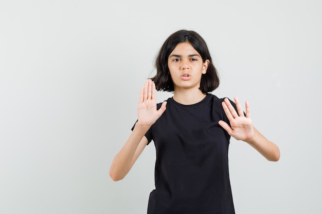 Little girl showing palms to defend herself in black t-shirt and looking careful. front view.