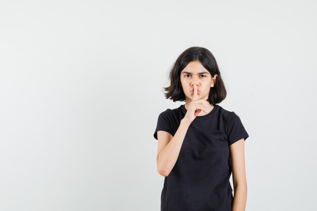 Little girl showing silence gesture in black t-shirt and looking confident , front view.