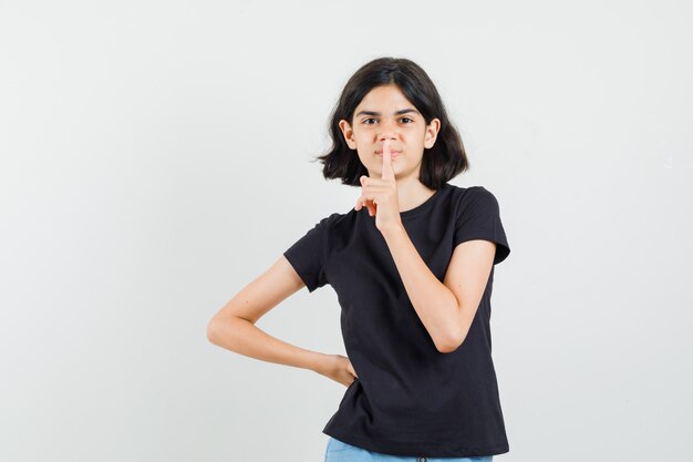 Little girl showing silence gesture in black t-shirt, shorts and looking careful. front view.