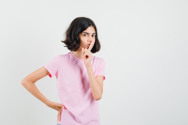 Little girl showing silence gesture in pink t-shirt and looking careful , front view.