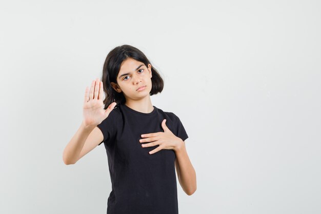 Маленькая девочка показывает жест остановки в черной футболке и выглядит усталой, вид спереди.