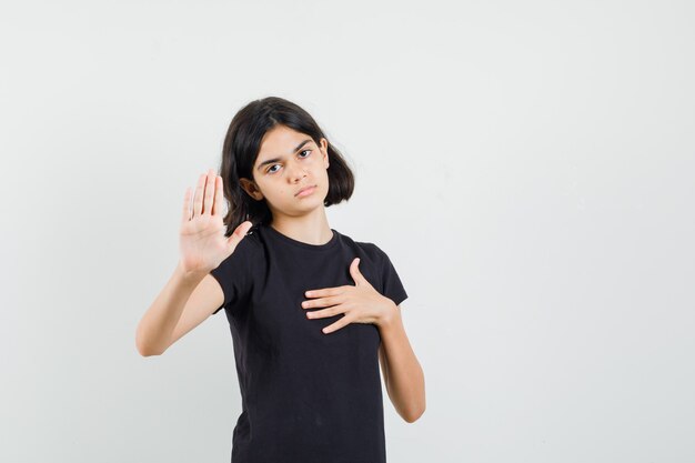 Little girl showing stop gesture in black t-shirt and looking tired , front view.