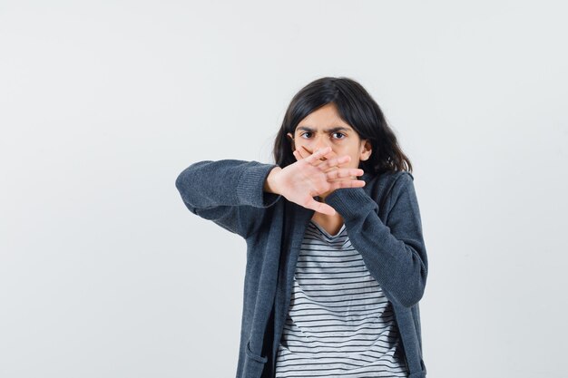 Little girl showing stop gesture, holding hand on mouth in t-shirt, jacket and looking scared.