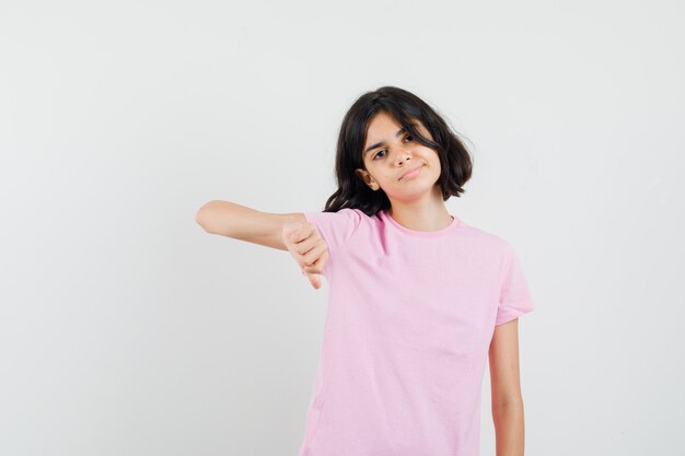 Little girl showing thumb down in pink t-shirt and looking confident , front view.