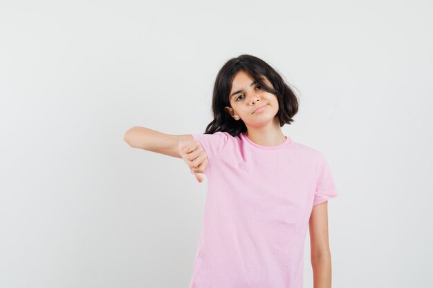 Маленькая девочка показывает палец вниз в розовой футболке и выглядит уверенно, вид спереди.