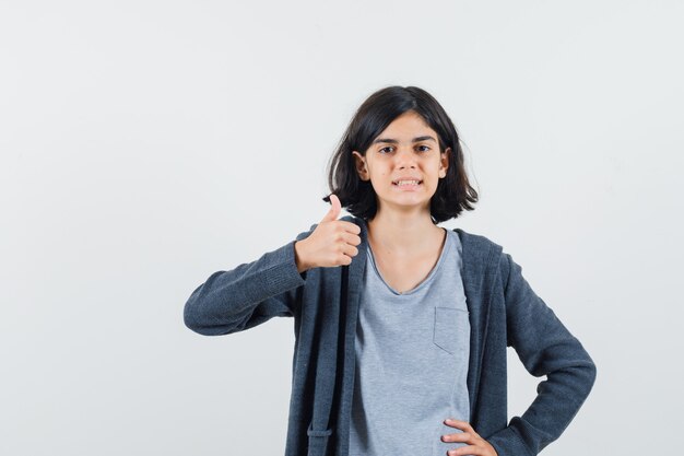 Little girl showing thumb up in t-shirt, jacket and looking pleased