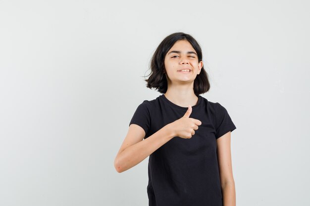 Маленькая девочка показывает палец вверх, подмигивая глазом в черной футболке, вид спереди.