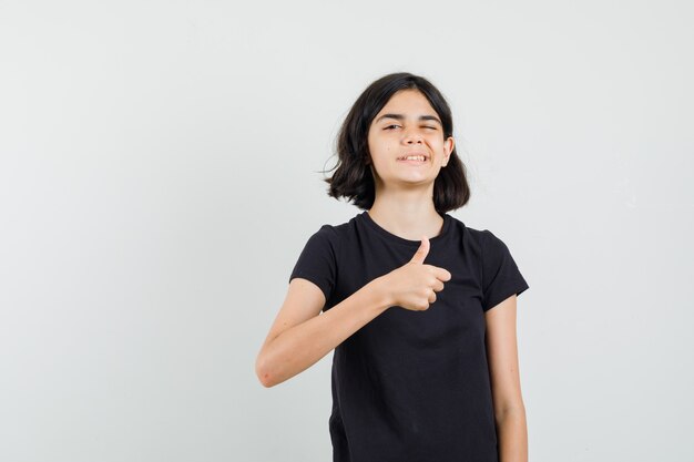 Little girl showing thumb up, winking eye in black t-shirt , front view.