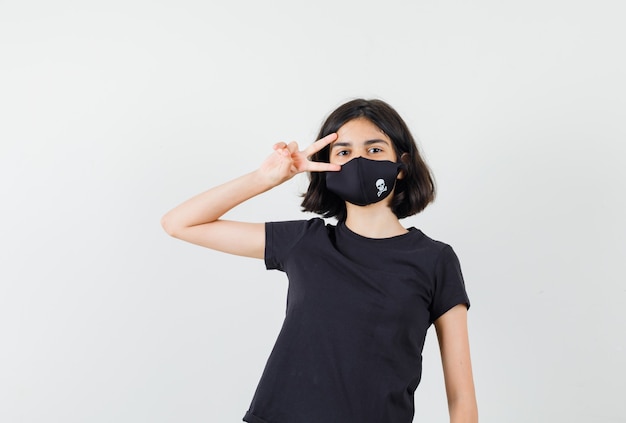 Little girl showing v-sign near eye in black t-shirt, mask front view.