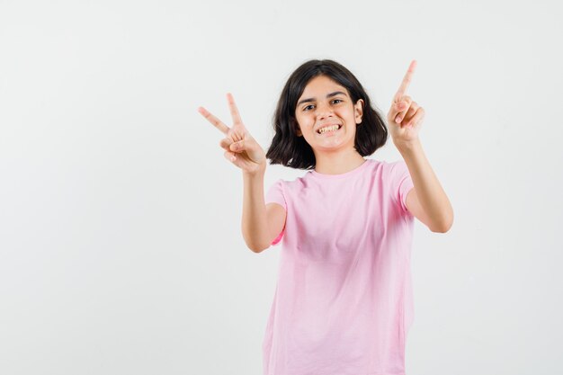 Little girl showing v-sign, pointing up in pink t-shirt and looking merry , front view.