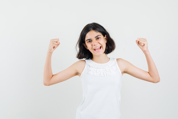 Little girl showing winner gesture in white blouse and looking lucky , front view.