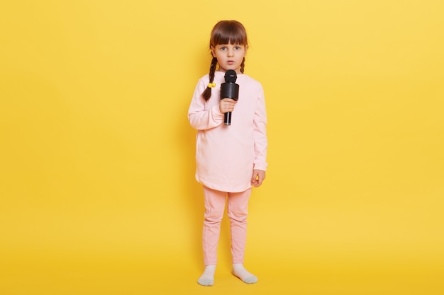Little girl singing song with cam and serious facial expression, looks at camera with worried look, being confused to arrange performance, wearing casual attire, isolated over yellow background.