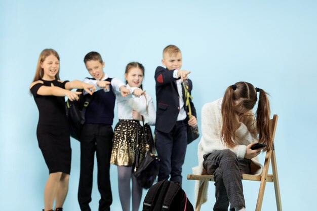 Little girl sitting alone on chair and suffering an act of bullying while children mocking. Sad young schoolgirl sitting on  against blue wall.