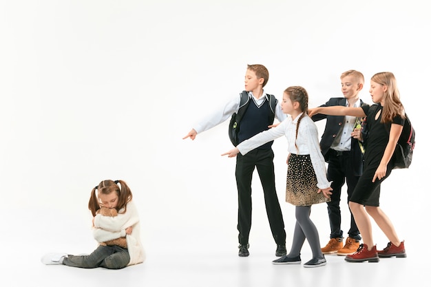 Little girl sitting alone on floor and suffering an act of bullying while children mocking. Sad young schoolgirl sitting on  against white wall.