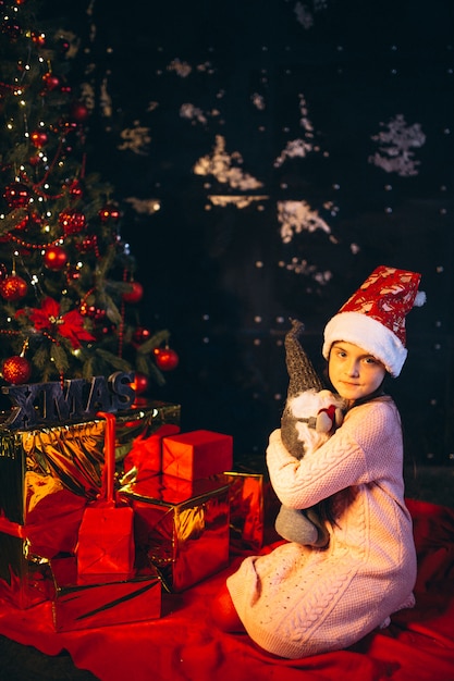 Маленькая девочка сидит возле елки и распаковывает подарки