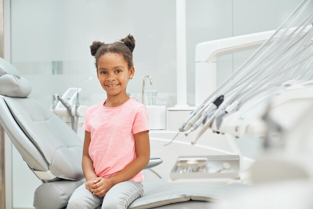 Маленькая девочка сидит на стоматологическом кресле и позирует в клинике