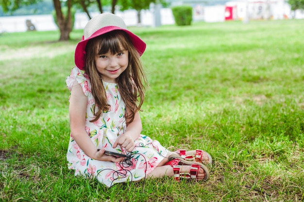 Маленькая девочка, сидя на траве, глядя на мобильный телефон с наушниками и в шляпе