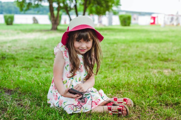 Маленькая девочка, сидя на траве, глядя на мобильный телефон с наушниками