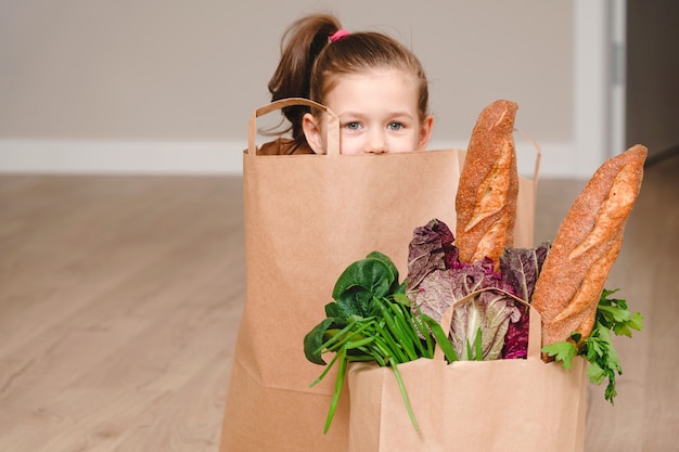 Маленькая девочка, сидя в бумажный мешок, прячась с овощами и хлебом, продуктовый с копией пространства