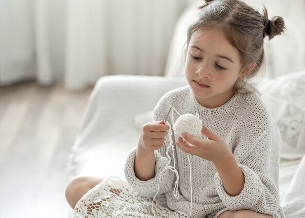 Little girl sitting on the sofa and learning to knit, home leisure concept.