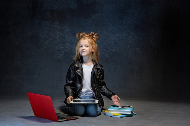 ノートパソコン、タブレット、灰色のスタジオで電話で座っている小さな女の子
