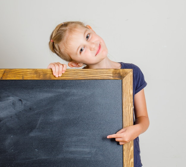 Little girl smiling and pointing at blackboard in t-shirt , front view.