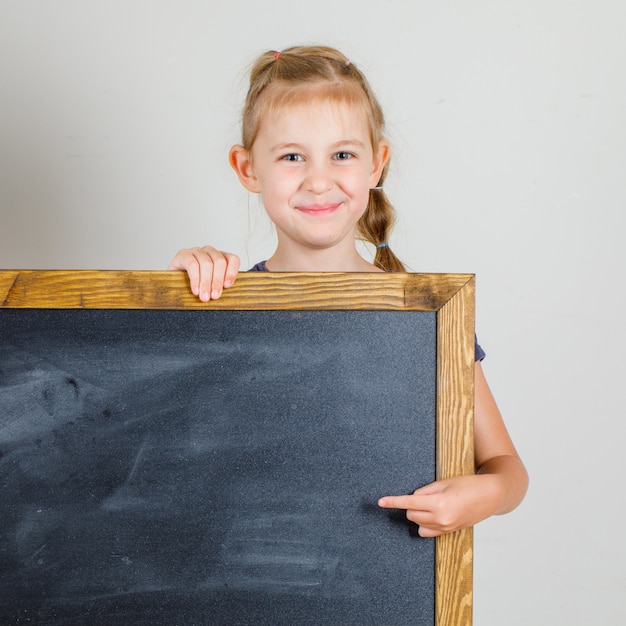 Free photo little girl smiling and pointing at blackboard in t-shirt front view.