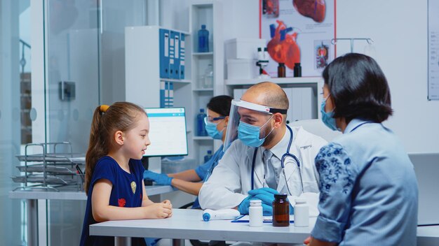 covid-19の相談中に医者と話している少女。病院のキャビネットで医療サービスの治療検査を提供する保護マスクを備えた医学の小児科医の専門家