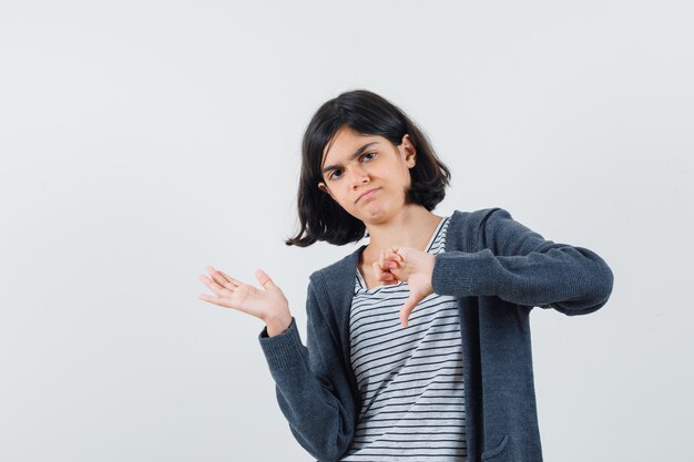 Little girl spreading palm aside, showing thumb down in t-shirt, jacket and looking discontent