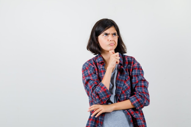 Little girl standing in thinking pose in checkered shirt and looking pensive , front view.