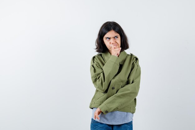 Little girl standing in thinking pose in coat, t-shirt, jeans and looking thoughtful , front view.