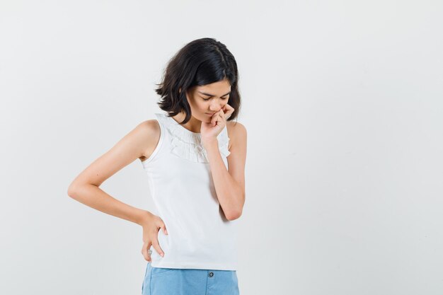 Little girl standing in thinking pose in white blouse, shorts front view.