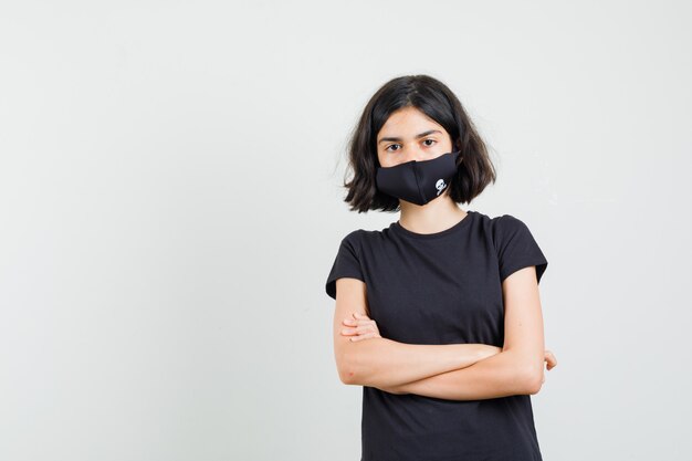 Little girl standing with crossed arms in black t-shirt, mask and looking sensible. front view.
