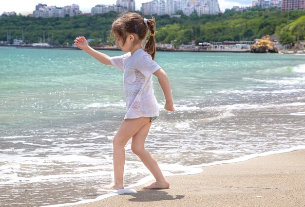A little girl stands barefoot on the seashore and wets her feet in the sea wave.