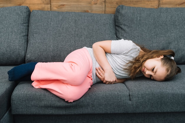 Little girl suffering from stomach ache lying on gray sofa