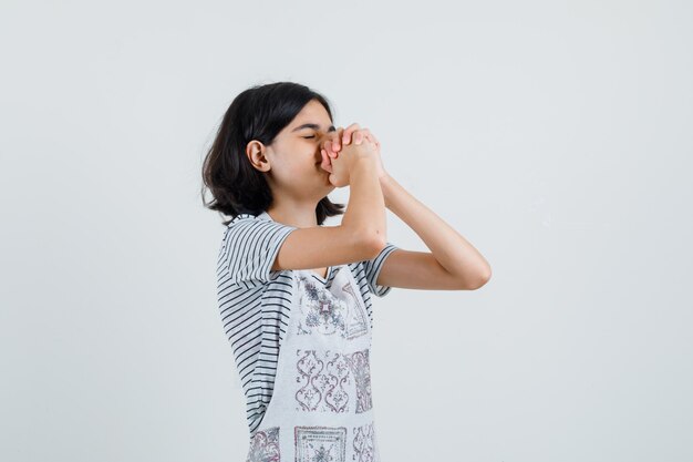 Little girl in t-shirt, apron clasping hands in praying gesture ,