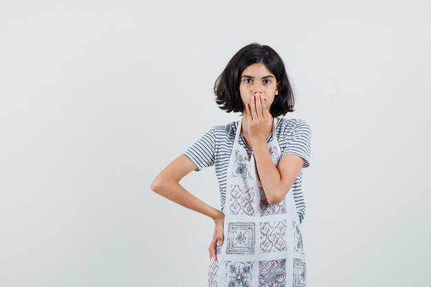 Little girl in t-shirt, apron holding hand on mouth and looking surprised ,