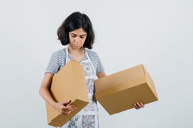 Little girl in t-shirt, apron looking at cardboard boxes and looking confused ,