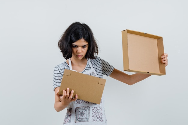 Free photo little girl in t-shirt, apron looking into cardboard box and looking puzzled ,