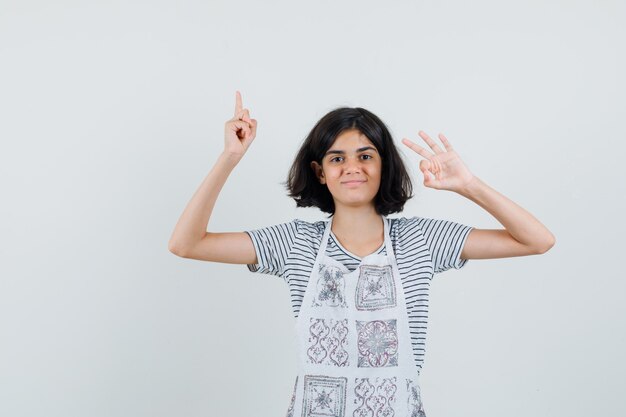 Little girl in t-shirt, apron pointing up, showing ok gesture and looking confident ,