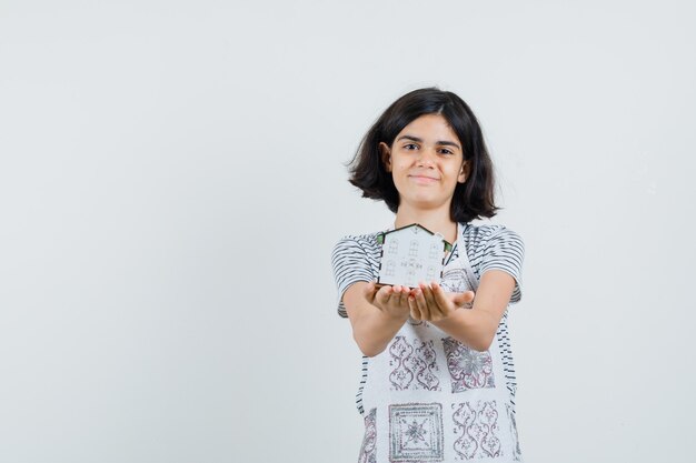 Little girl in t-shirt, apron presenting house model and looking cheerful ,