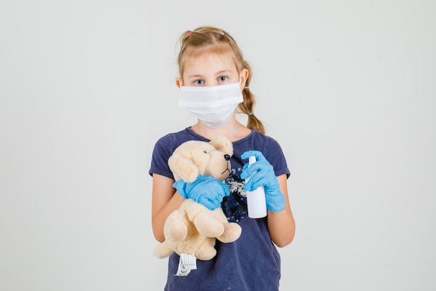 Little girl in t-shirt, gloves and medical mask desenfecting toy bear , front view.