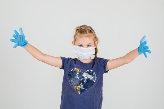 Little girl in t-shirt, gloves and medical mask raising her palms up and looking happy , front view.